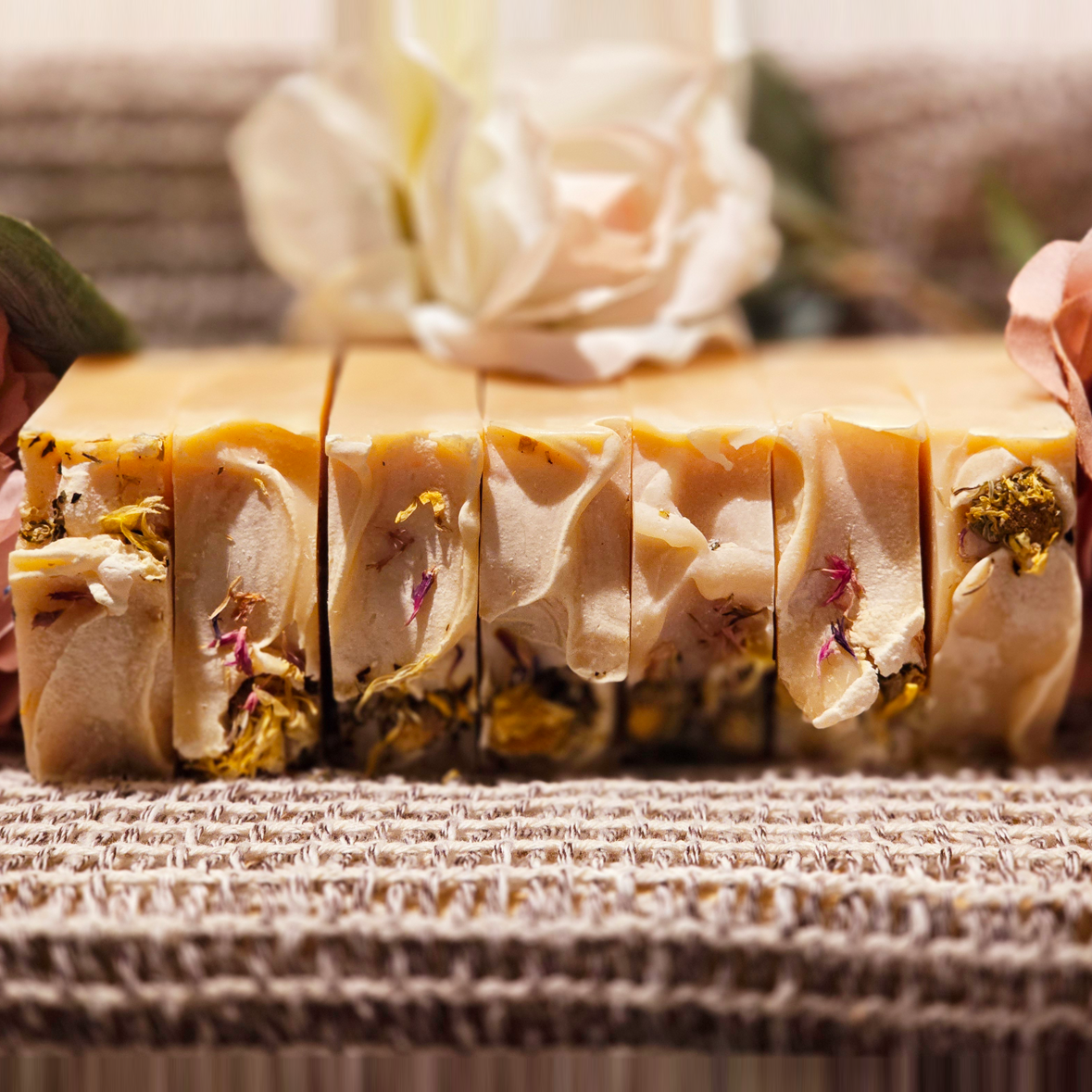 Row of Secret Garden soap bars lined up, showcasing creamy swirls, dried botanicals, and vibrant flower accents against a woven textured backdrop with roses.