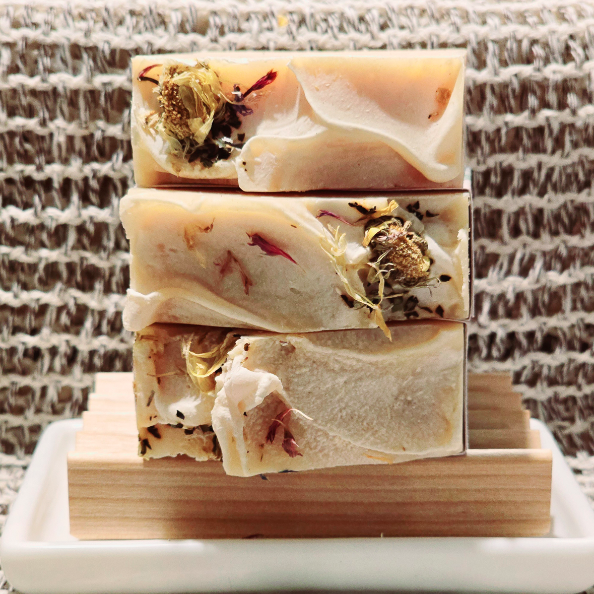 Three Secret Garden soap bars stacked, showing carrot-hued swirls, dried flowers, and creamy textures, placed on a wooden soap dish against a woven backdrop.