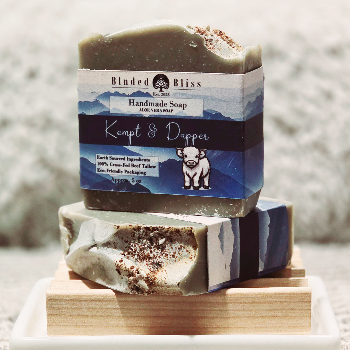 Kempt & Dapper handmade aloe vera soap with a deep green hue, natural coconut shell powder topping, and sleek blue mountain-inspired label, stacked on a wooden soap dish.