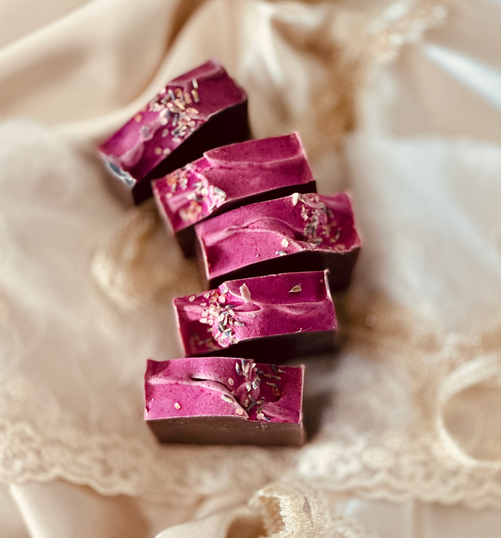 A close-up of five lavender soap bars with a smooth, purple-tinted top layer and a dark base, adorned with dried lavender buds